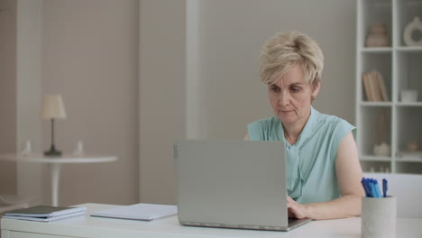 Una-Mujer-Jubilada-Está-Escribiendo-En-Una-Computadora-Portátil.-Una-Escritora-Está-Trabajando-Y-Escribiendo-En-Casa-Mirando-La-Pantalla-Con-Concentración.